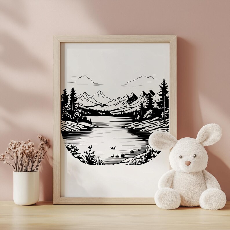 May include: Black and white framed art print of a mountain lake scene. The artwork depicts a serene landscape with mountains, trees, and a lake. The frame is light-colored, and the print is displayed on a wooden surface.