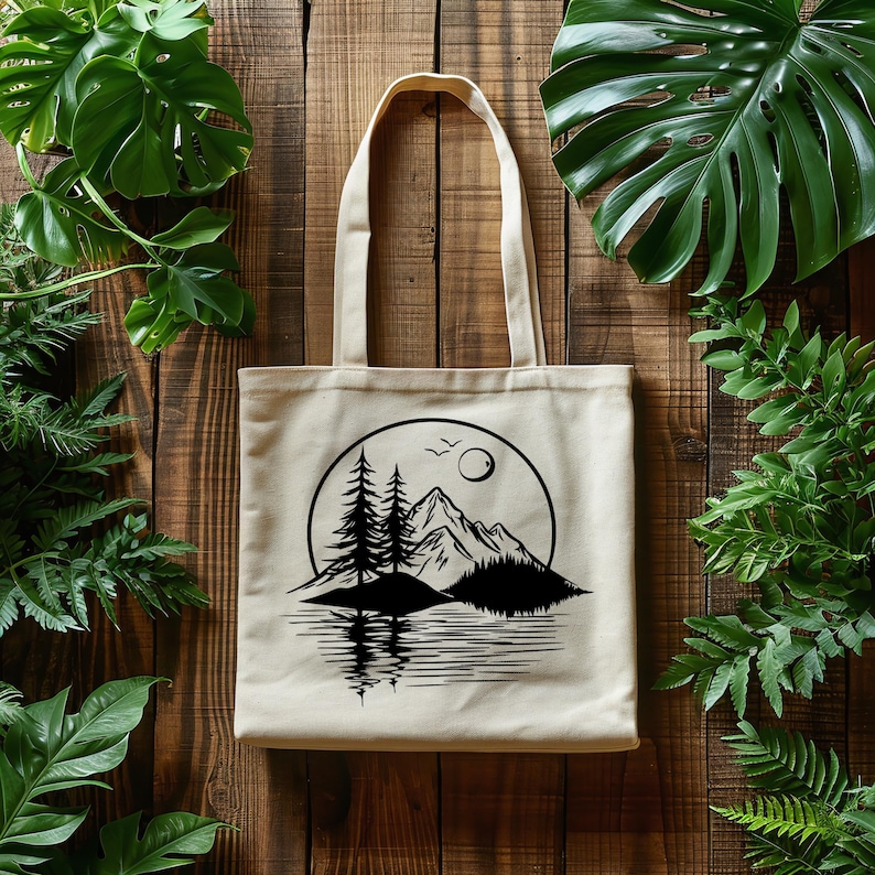 May include: Beige canvas tote bag featuring a black and white mountain and lake scene within a circle. The design includes pine trees, mountains, a sun, and birds. The bag has a rectangular shape with two handles.