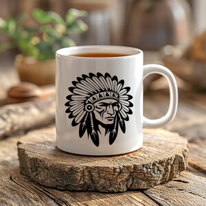 May include: White ceramic mug featuring a black graphic of a Native American chief with a headdress. The mug is filled with a dark liquid and sits on a wooden surface. The mug has a curved handle.