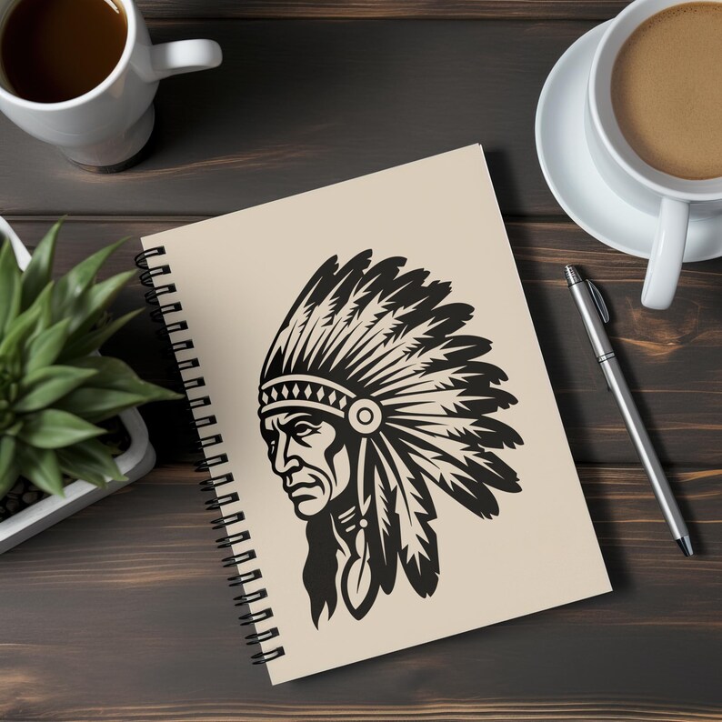 May include: Spiral-bound notebook featuring a black and white illustration of a Native American chief in profile. The notebook has a cream-colored cover and is resting on a dark wooden surface, alongside coffee cups and a pen.
