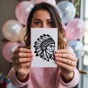 May include: A white card with a pink border featuring a black and white illustration of a Native American chief in profile. The chief is wearing a feathered headdress. The card is held in front of a person, with balloons in the background.