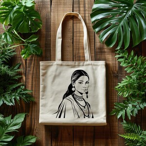 May include: Beige tote bag featuring a black and white illustration of a woman with long hair, earrings, and necklaces. The tote bag has long handles and is made of a canvas-like material. The background is a wooden surface with green plants.