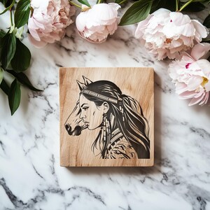 May include: A square wooden plaque with a detailed black and white illustration of a woman's profile merged with a horse's head. The woman wears a headband and long earrings. The plaque is set against a marble surface with pink peonies.