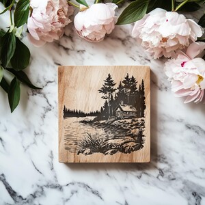 May include: A square wooden cutting board with a black etched landscape scene featuring a cabin, trees, and a lake. The board is surrounded by pink peonies and green leaves on a marble surface.