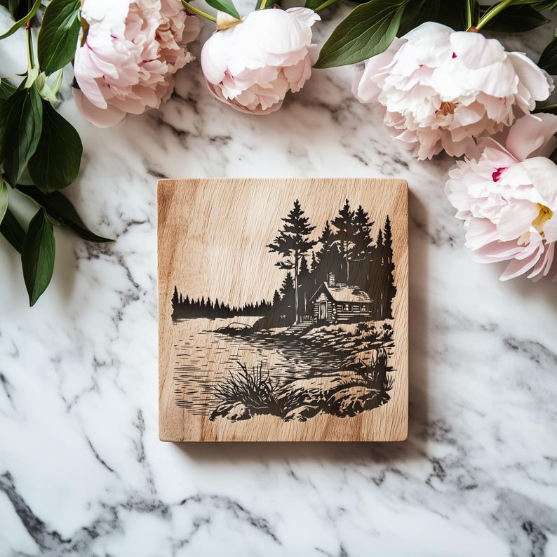 May include: A square wooden cutting board with a black etched landscape scene featuring a cabin, trees, and a lake. The board is surrounded by pink peonies and green leaves on a marble surface.