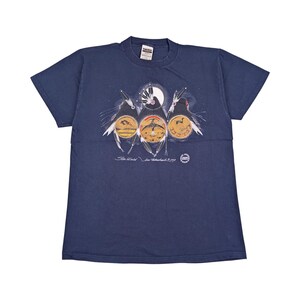 Peut inclure: T-shirt bleu marine avec un motif de trois oiseaux stylisés, chacun avec un motif circulaire sur la poitrine, sur fond de ciel nocturne avec une pleine lune. Le t-shirt comporte du texte en bas et un petit logo à droite.