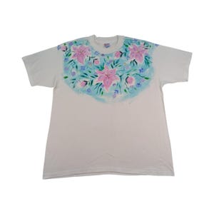 May include: White t-shirt with a hand-painted floral design. The design features large pink lilies, small blue flowers, and green leaves on a light blue background. The shirt is a classic crew neck style.