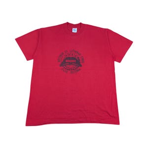 Peut inclure: Un t-shirt rouge à col rond et à manches courtes. Le t-shirt présente un motif graphique gris foncé sur le devant avec le texte "Wisconsin Ev. Lutheran Synod Apache Centennial".