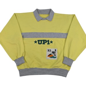 Könnte beinhalten: Gelber und grauer Pullover mit einem grauen Streifen über der Brust und einem grünen gestickten Logo, das "UP1" mit Sternen auf beiden Seiten sagt. Das Logo befindet sich über einem kleinen rechteckigen Patch mit dem Text "K1" und einem Cartoon-Bild einer Person auf einem Surfbrett.