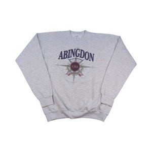 Op de afbeelding: Een lichtgrijs sweatshirt met het woord "ABINGDON" in marineblauw gebogen bovenaan. Daaronder staat het woord "ILLINOIS" in kleinere letters. Een kompasontwerp met het woord "USA" in het midden.