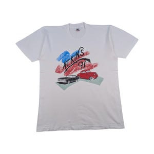 Könnte beinhalten: Weißes T-Shirt mit Grafikdruck. Das Design zeigt den Text "ATKINS 91", ein schwarzes Auto und ein rotes Auto, vor blau-roten Pinselstrichen.