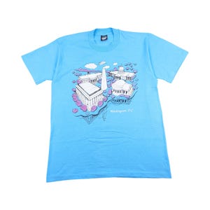 Könnte beinhalten: Hellblaues T-Shirt mit einem Grafikdruck von Wahrzeichen von Washington, D.C., darunter das Washington Monument und das Capitol Building. Der Text "Washington, DC" ist darunter gedruckt. Das Shirt ist aus weichem Material.