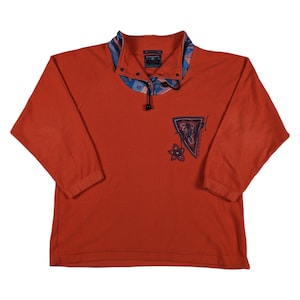 Puede incluir: Sudadera de forro polar naranja con cremallera negra y cuello de estampado colorido. La parte delantera de la sudadera presenta un diseño gráfico con una flor y un triángulo.