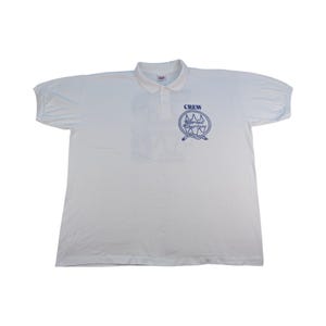 Könnte beinhalten: Weißes Poloshirt mit kurzen Ärmeln. Das Shirt hat eine blaue Grafik auf der Brust mit der Aufschrift "CREW" über einem nautischen Design und den Worten "Sailor Tail Charters". Das Shirt hat einen Kragen.