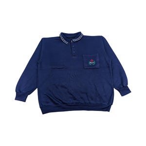 Könnte beinhalten: Ein marineblaues Langarm-Sweatshirt mit Kragen und einer kleinen Tasche auf der Brust. Der Kragen hat ein weißes und silbernes kettenartiges Design. Die Tasche zeigt ein rot-grünes Emblem mit dem Wort "Royal".