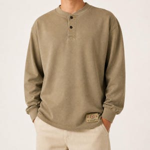 Könnte beinhalten: Olivgrünes Langarmshirt mit Henley-Ausschnitt und drei dunklen Knöpfen. Das Shirt hat eine lockere Passform und einen kleinen Aufnäher am Saum. Das Shirt wird mit einer khakifarbenen Hose kombiniert.