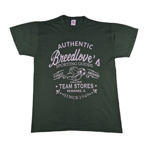 Könnte beinhalten: Ein dunkelgrünes T-Shirt mit einem weißen Grafikdruck, auf dem "Authentic Breedlove's Sporting Goods" steht, mit einem Design aus Sportbällen und dem Text "Finest Online Team Stores Kewanee, IL Since 1949."
