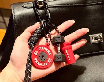 May include: A fitness-themed keychain featuring a red weight plate with "Fitness" text and 45 LB markings, a dumbbell with 5 KG markings, and a red sport bottle. The keychain is attached to a black and white rope.