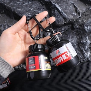 May include: Two black plastic containers with labels that say "Gold Standard 100% Whey" and "Whey" are attached to black carabiner clips. The containers are for protein powder or supplements.