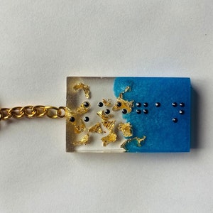 May include: A blue and white resin keychain with gold flakes and black dots. The keychain is shaped like a rectangle and has a gold chain attached to it.