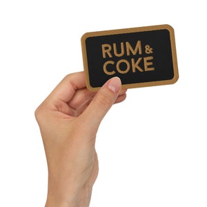 Op de afbeelding: Een rechthoekige bruine en zwarte patch met de tekst "RUM & COKE" in gouden borduurwerk.