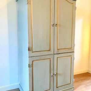 May include: A tall, light blue cabinet with four doors. The cabinet has gold trim around the doors and edges, with decorative hardware. The top of the cabinet has a curved design.