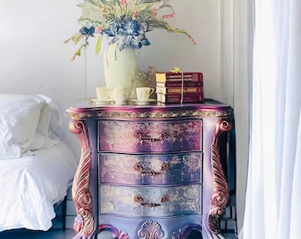 May include: A purple and gold painted nightstand with three drawers and ornate carvings. The nightstand has a white marble top with a bouquet of purple flowers in a white vase and a stack of books on top.