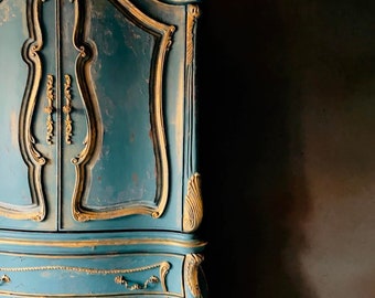 May include: A tall, ornate armoire with a teal blue finish and gold accents. The armoire has two doors with intricate carvings and three drawers below.