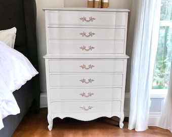 May include: A white, antique-style dresser with six drawers and ornate rose gold handles. The dresser is placed in a bedroom setting, with a bed and window visible in the background. The dresser is approximately 50 inches tall.