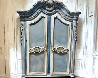 May include: A large, antique, two-door armoire with a carved, ornate top. The armoire is painted in a light blue and gold color scheme. The doors are closed and have a decorative, gold trim. The armoire is standing on a wooden floor.