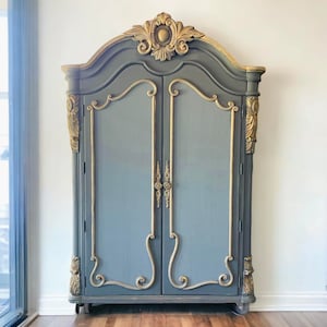 May include: A tall, gray armoire with gold accents and ornate carvings. The armoire has two doors and sits on wheels.
