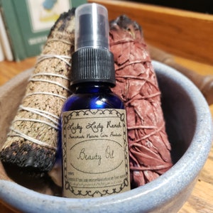 May include: A blue glass bottle with a spray nozzle and a brown paper label that reads "Lucky Lady Ranch Homemade Nature Care Products Beauty Oil". The bottle is sitting in a blue ceramic bowl with two bundles of dried sage.