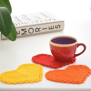 Op de afbeelding: Drie gehaakte hartvormige onderzetters in geel, oranje en rood. De onderzetters liggen op een wit oppervlak met een boek en een kop koffie.