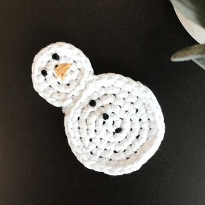 May include: A handmade white crochet snowman with black button eyes and a small orange nose. The snowman is made of two connected circles, one larger for the body and a smaller one for the head. The snowman is on a black surface.