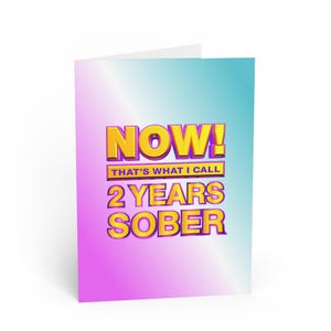 May include: A greeting card with a gradient background of pink, blue, and white. The card features the text "NOW! THAT'S WHAT I CALL 2 YEARS SOBER" in a bold, yellow, 3D font with a purple outline.