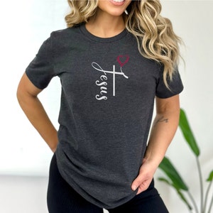 K&ouml;nnte beinhalten: Ein dunkelgraues T-Shirt mit einem wei&szlig;en Kreuz und dem Wort "Jesus" in Kursivschrift. Ein rotes Herz befindet sich &uuml;ber dem Kreuz.