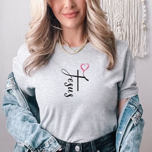 K&ouml;nnte beinhalten: Ein graues T-Shirt mit einem schwarzen Kreuz und dem Wort "Jesus", das vertikal daneben geschrieben ist. Ein rosa Herz befindet sich &uuml;ber dem Kreuz.