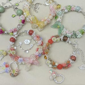 May include: A collection of floral bracelets and rings in various colors, including pink, white, yellow, and green. The bracelets feature delicate flower and bead accents, with silver-toned chains and charms. Some bracelets have a butterfly or bow design.