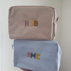 May include: Two rectangular cosmetic bags. The top bag is tan and white striped with the embroidered letters "HUB" in pastel colors. The bottom bag is blue and white striped with the embroidered letters "AMC" in pastel colors.