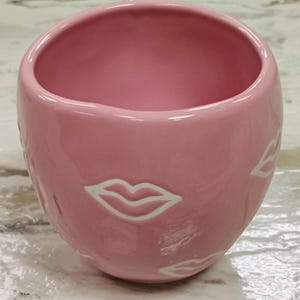 May include: A pink ceramic mug with a glossy finish. The mug features a rounded shape and is adorned with white lip designs around the exterior. The interior of the mug is also pink.