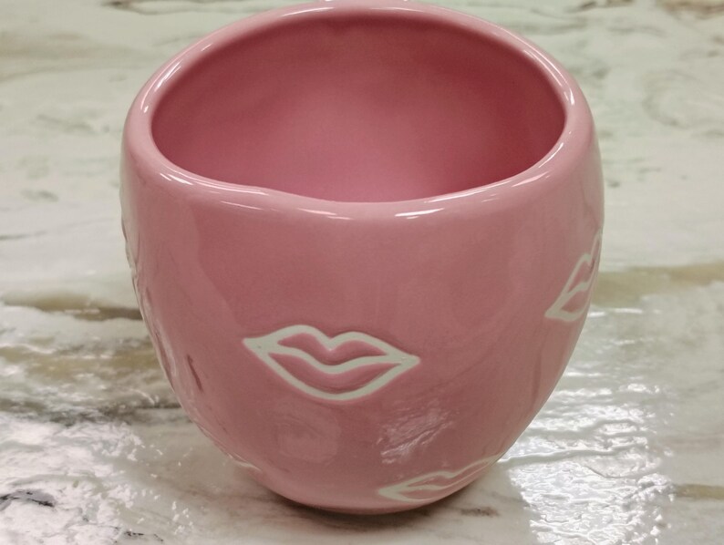 May include: A pink ceramic mug with a glossy finish. The mug features a rounded shape and is adorned with white lip designs around the exterior. The interior of the mug is also pink.