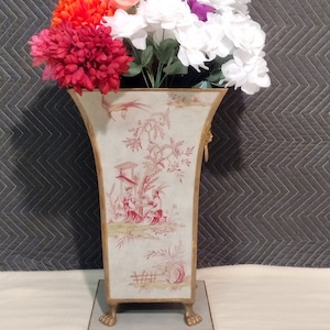 May include: A white ceramic vase with gold accents and a floral design. The vase has three gold feet and is filled with a bouquet of artificial flowers in shades of red, pink, and white.