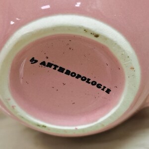 May include: Close-up of a pink ceramic mug with a white rim. The text "by ANTHROPOLOGIE" is printed in black on the bottom of the mug. The mug has a glossy finish and a simple, elegant design.