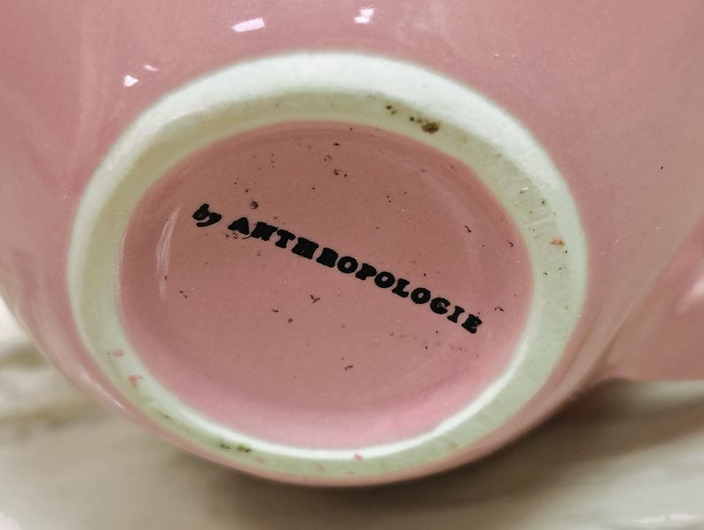 May include: Close-up of a pink ceramic mug with a white rim. The text "by ANTHROPOLOGIE" is printed in black on the bottom of the mug. The mug has a glossy finish and a simple, elegant design.