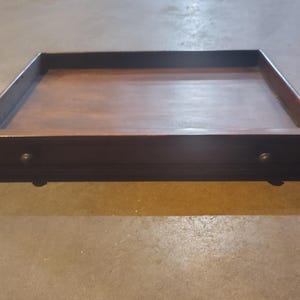 May include: A rectangular wooden tray with a dark brown finish. The tray has a raised edge and small, round metal knobs on the front. The tray is designed to sit on four small feet.