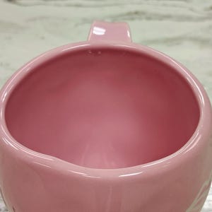May include: A close-up of a pink ceramic mug with a handle. The mug has a smooth, glossy finish and a rounded shape. The interior of the mug is also pink. The mug is likely used for drinking beverages.