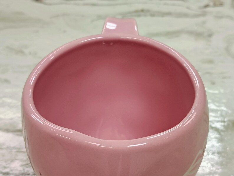 May include: A close-up of a pink ceramic mug with a handle. The mug has a smooth, glossy finish and a rounded shape. The interior of the mug is also pink. The mug is likely used for drinking beverages.