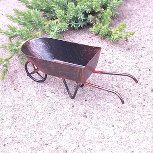 May include: A miniature metal wheelbarrow with a distressed, reddish-brown finish and a black interior. The wheelbarrow has a single wheel and two handles. The background features green foliage and a textured gray surface.