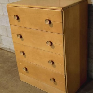 May include: A light-colored wooden dresser with five drawers and round wooden knobs. The dresser has a simple, classic design and is set against a neutral-colored wall.