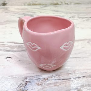 May include: A pink ceramic mug with a handle, featuring a face design with white lips. The mug has a smooth, glossy finish and is set against a marble-like surface. The mug is designed for beverages.